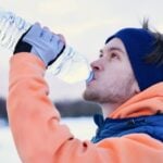 Jovem com sede está andando e bebendo água pura e fria no dia de neve no inverno.