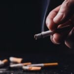 Pessoa fumando, bitucas de cigarro esoalhadas ao fundo. Imagem com fundo preto.