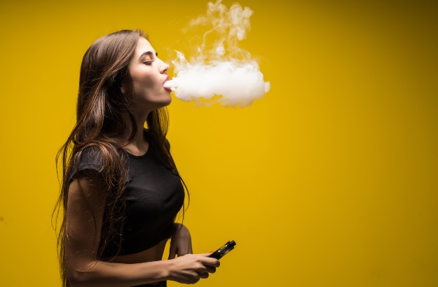 Jovem usando vape, soltando fumaça e com fundo amarelo