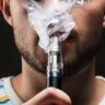 Homem de barba rala usando vape, fundo preto