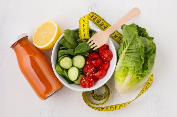 Uma dieta saudável composta por legumes, frutas, suco natural, verduras em prato branco e fundo branco com fita métrica